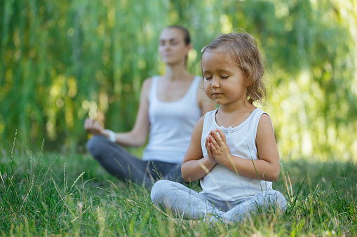 Young mother and little girl meditating in lotus pose together at green summer park kids meditation