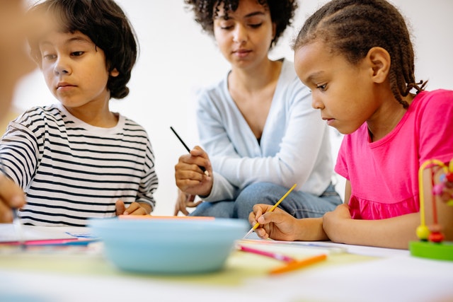 Woman sitting with kids at a table painting What is taught to kids in kindergarten
