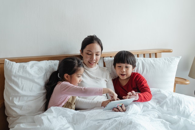 Woman and kids sitting on bed while playing on tablet