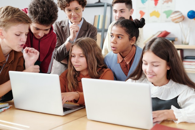 Two girls using laptop with classmates as teacher stands behind