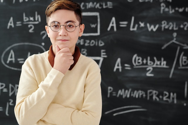 Young boy wearing glasses standing in front of blackboard