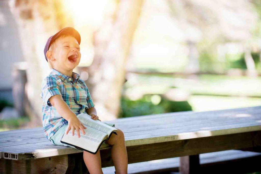 Child sitting on a bench table, holding an open book and laughing