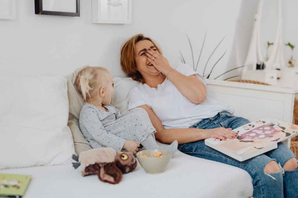 A woman and a child sitting on a cot laughing as they read a book