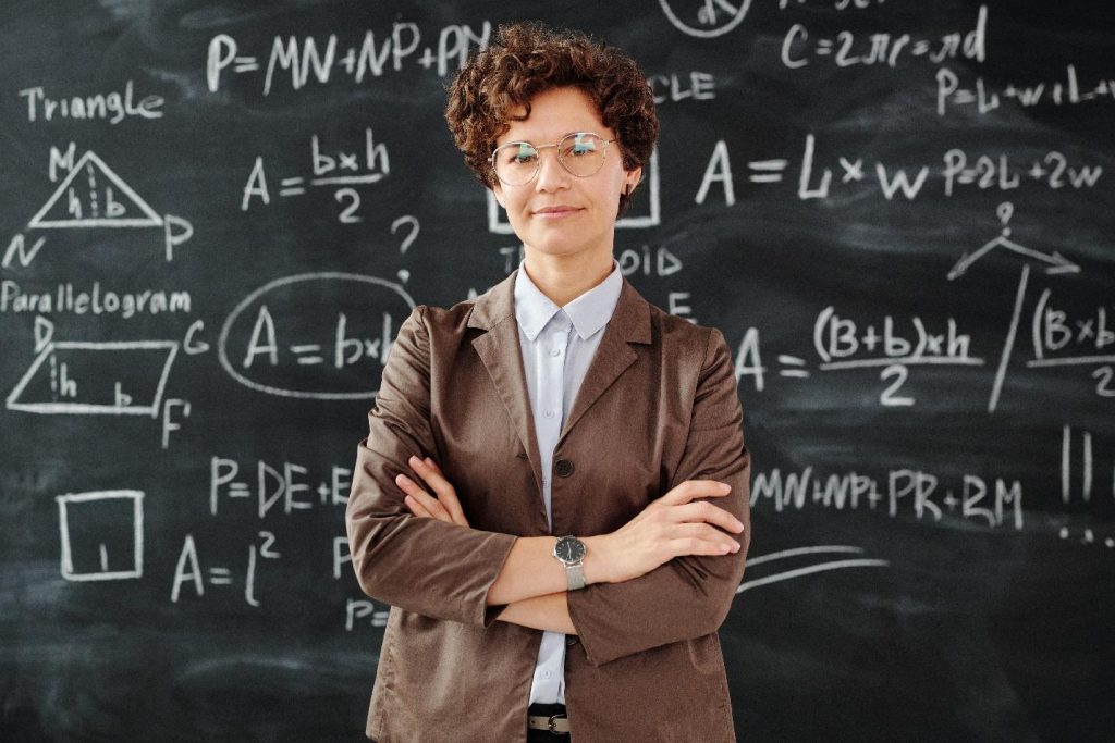 howdidnewcommoncoremathoriginate Teacher with arms crossed standing in front of blackboard