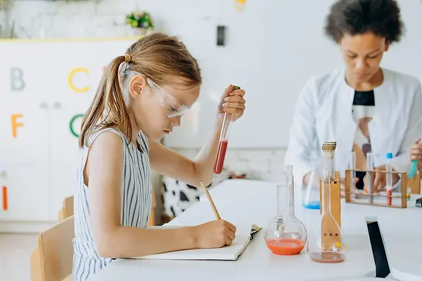 Young girl holding a test tube and writing in a notebook the teacher is in the background