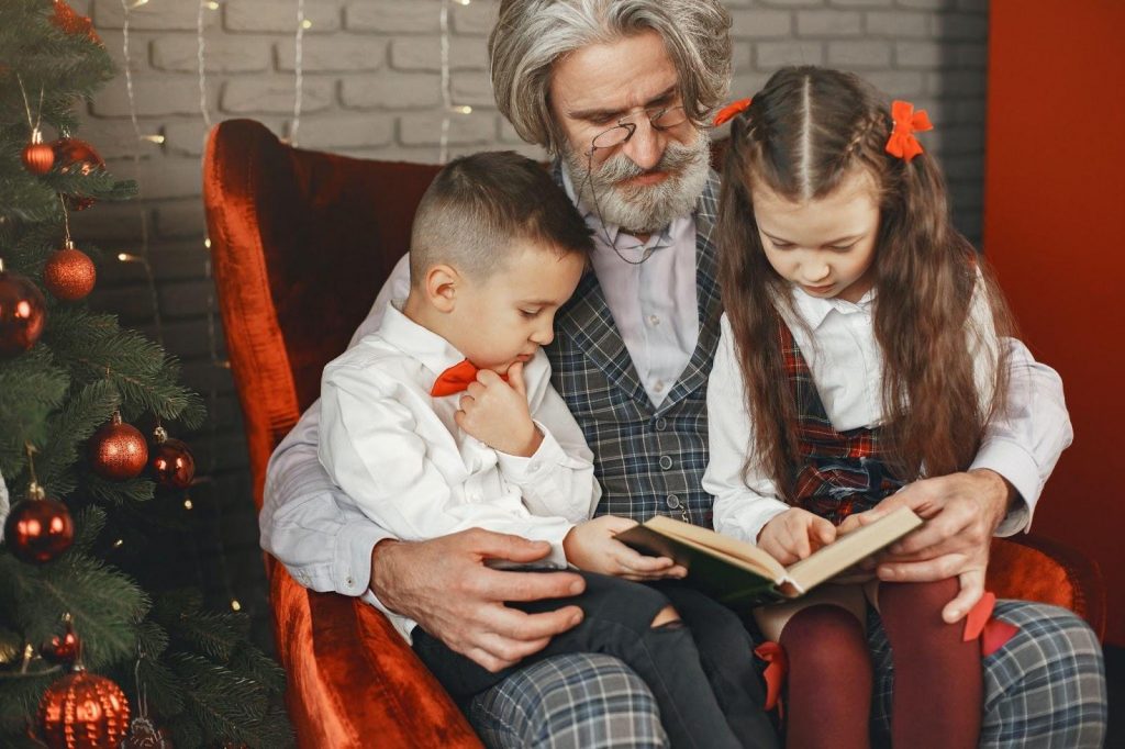 Grandfather holding his grandchildren on his lap and reading to them next to a Christmas tree