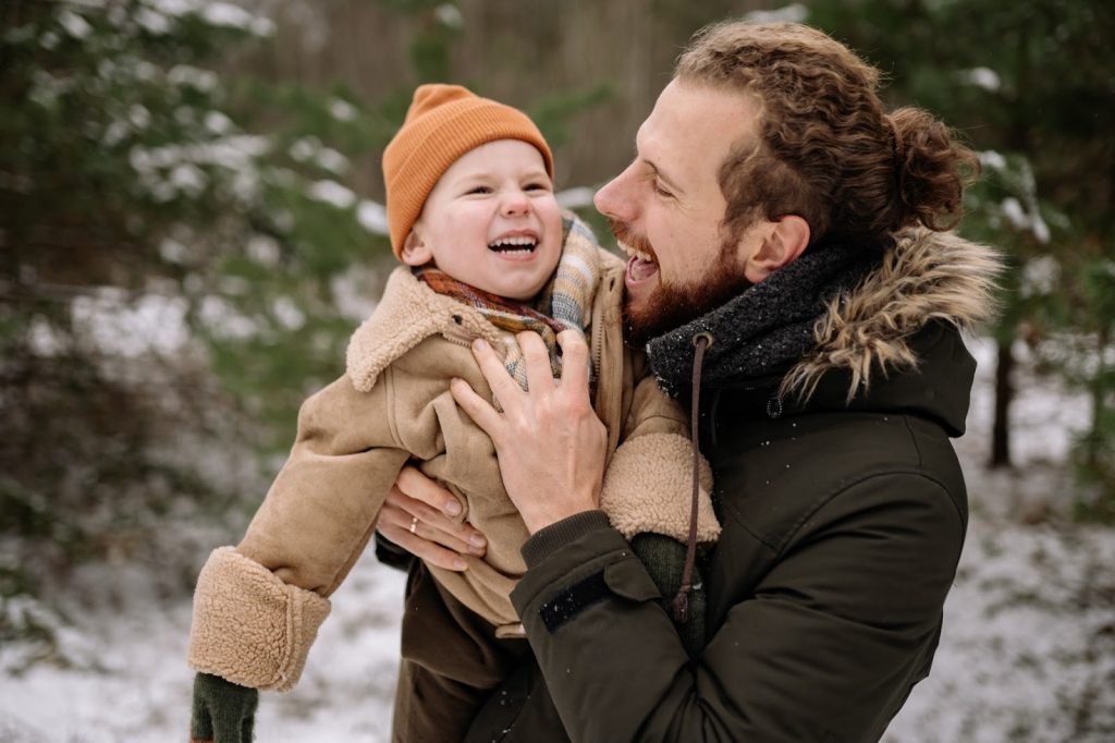 A father holds a child outdoors in the snow as they laugh together