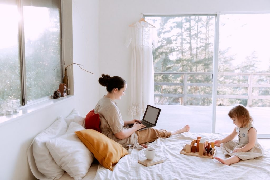 Mother working on computer and child playing with wooden toys on a bed