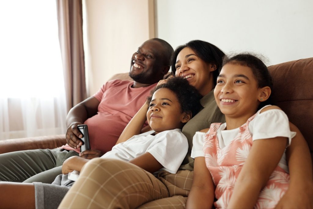 A family of four smiles as they share a couch and watch TV