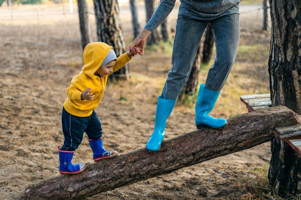 A toddler walks up an elevated log with a parents support