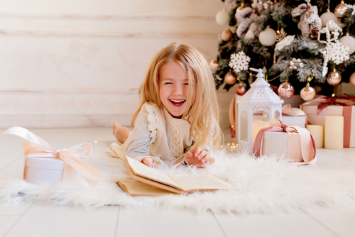 Little girl reading a book and laughing in front of a Christmas tree