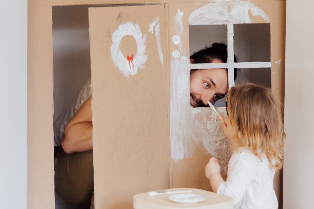 Girl paints her fathers nose through the window of cardboard house