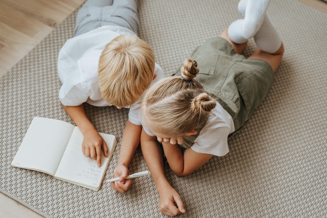 Two kids laying on a mat writing in a notebook