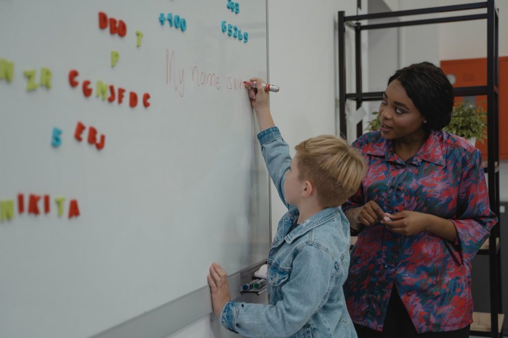 A young boy writes on a whiteboard as a teacher watches from the side