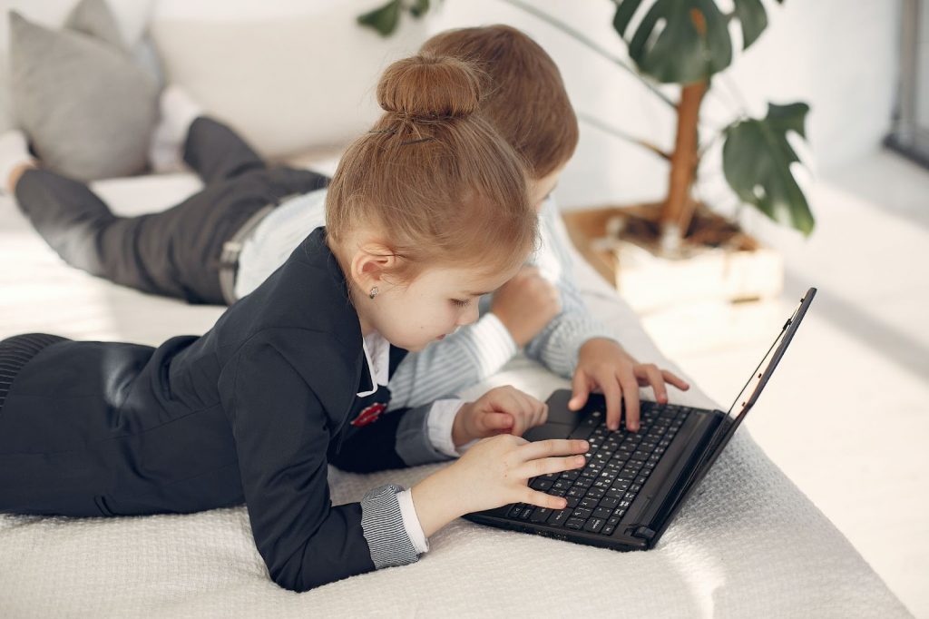 Two kids lying on bed and playing on laptop