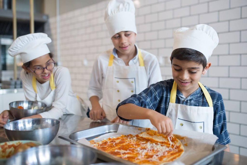 Young kid in plaid shirt making a pizza while standing near his friends