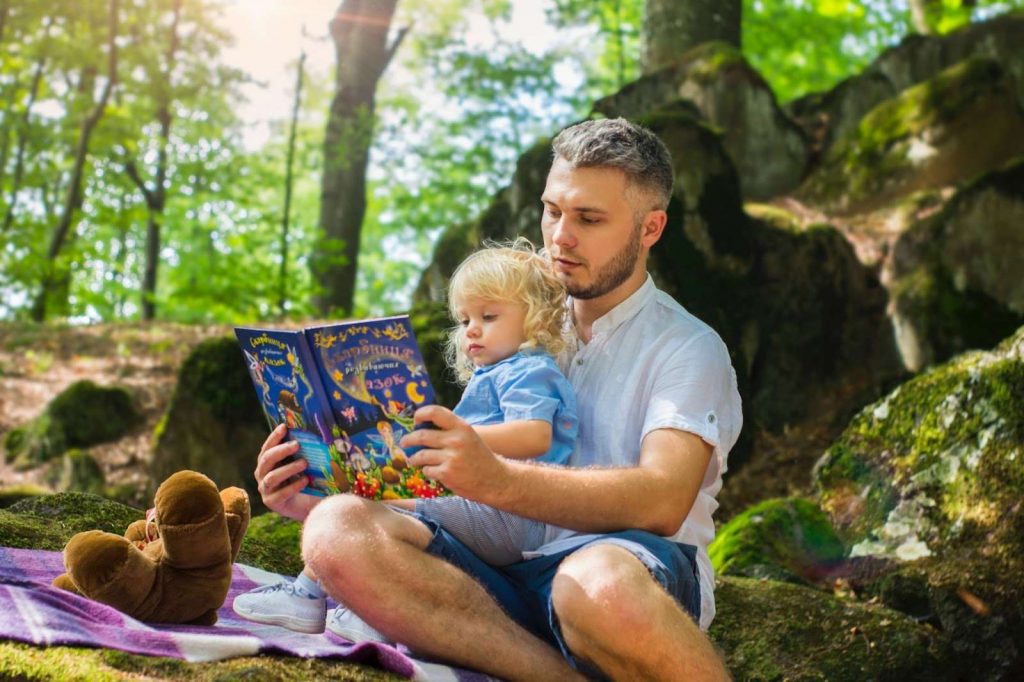 Father reading a book with her toddler in the lap of nature